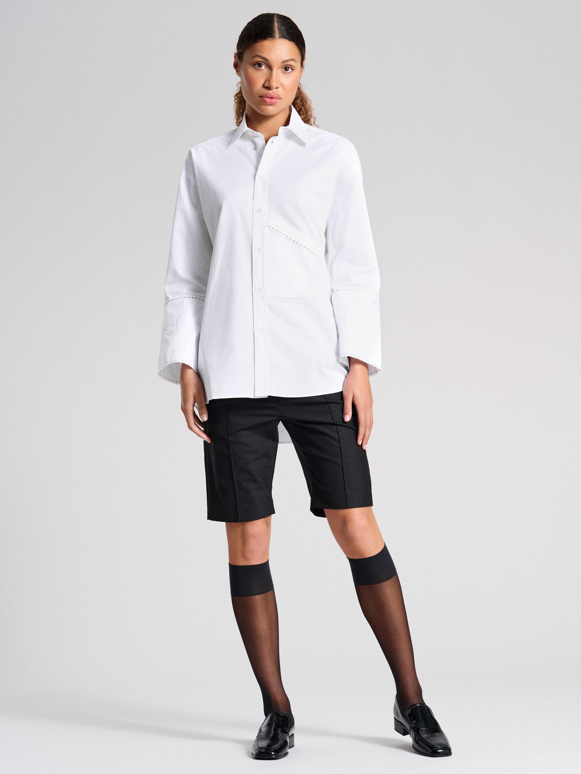 Chic office look with model wearing black knee-high socks paired with a crisp white shirt, Bermuda shorts, and classic black loafers.