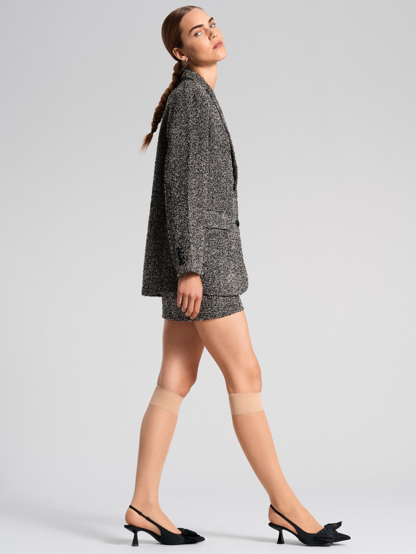 A model strides in nude knee-high socks, teamed with a textured grey blazer and shorts, and black slingback heels, embodying a modern, sophisticated style.
